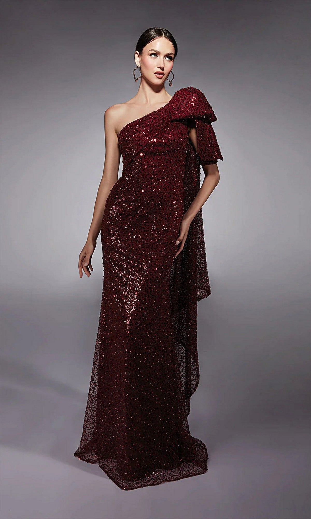 A woman wearing the Alyce 27771 long formal dress in wine, featuring a floor-length one-shoulder sequin design with a large bow detail, stands against a plain gray background.